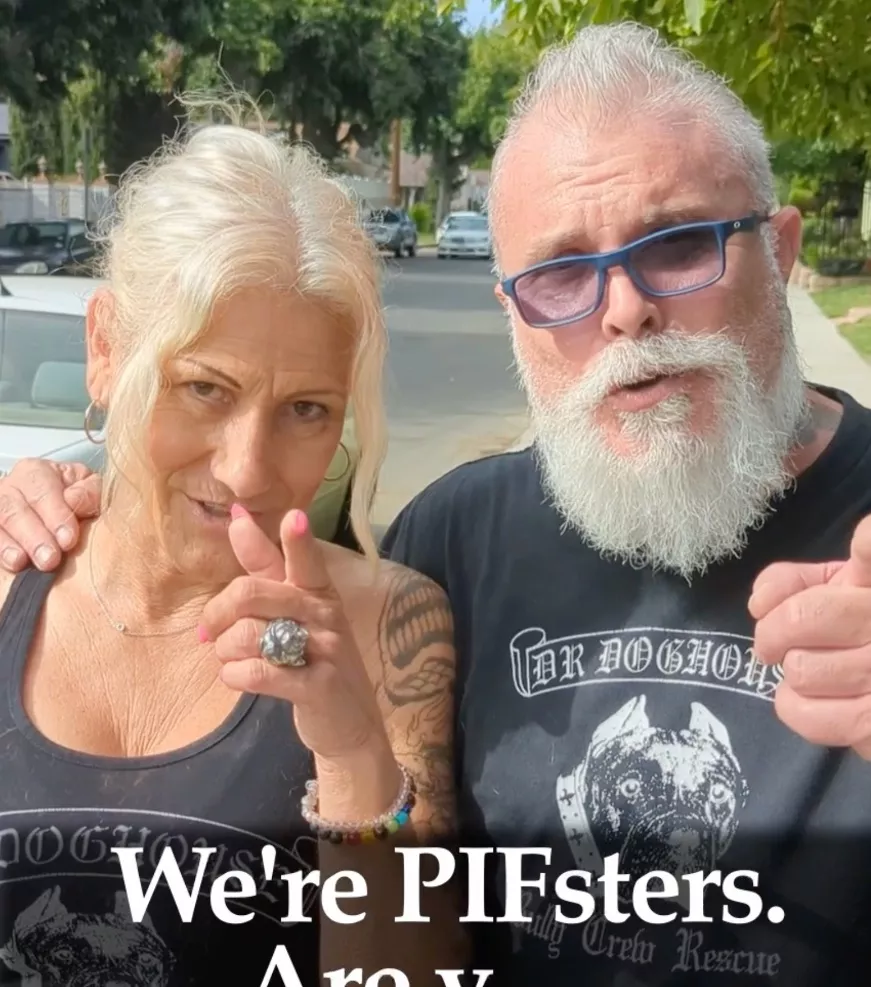 An older woman and man with tattoos and white hair stand outdoors, pointing at the camera and smiling. Both wear black Dr Doghouse shirts, showing their spirit of giving. White text at the bottom reads, Were PIFsters. Are you.