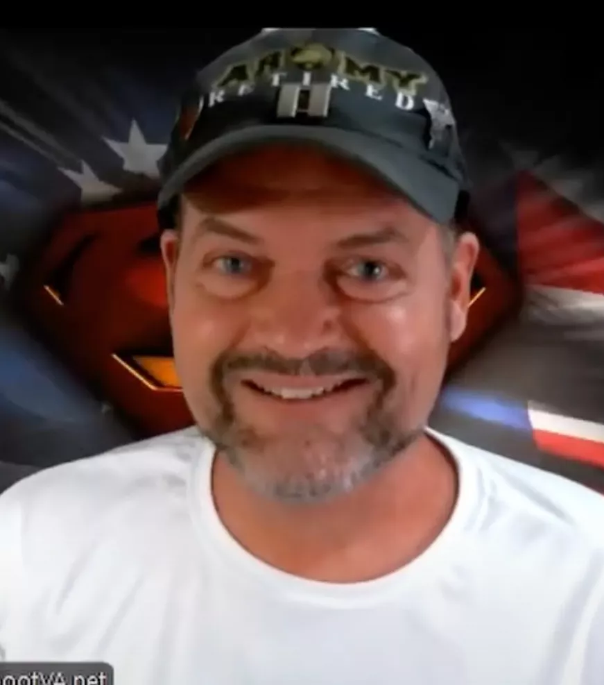 A smiling man with a beard wears a dark cap and a white shirt. Behind him, a backdrop featuring a stylized Superman logo and elements of the American flag highlights his commitment to giving and help.
