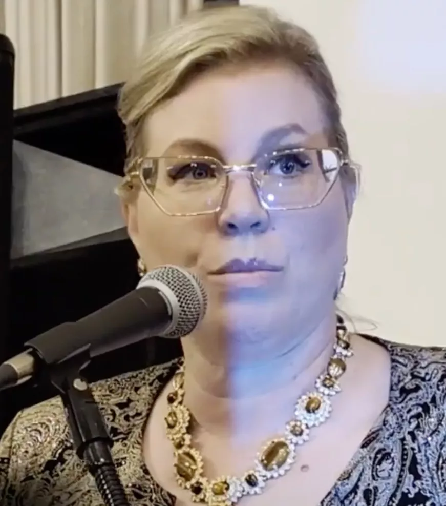 A woman with blond hair, wearing glasses and a decorative necklace, speaks into a microphone about giving. She is dressed in a patterned top, and the lighting casts a distinct vertical shadow across her face.