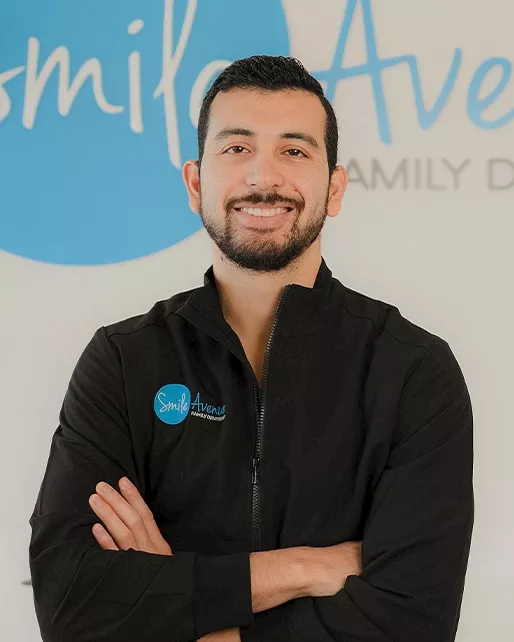 Dentist at Smile Avenue Family Dentistry standing in front of the clinic's logo in Katy, TX. Expert in family, emergency, and cosmetic dental care, wearing a black uniform with the clinic's logo.
