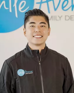 Smiling male dentist wearing a black jacket with the Smile Avenue Family Dentistry logo in front of a branded background at Cypress TX Family Dentist & Cosmetic Care office.