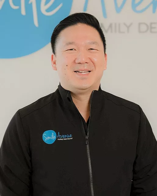 A dentist wearing a black Smile Avenue branded jacket is smiling in front of a Smile Avenue Family Dentistry sign, representing dental care services in Katy, TX.