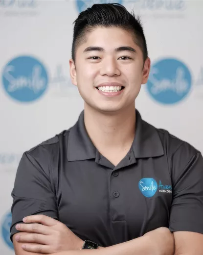 A smiling dentist with dark hair wearing a gray polo shirt featuring the Smile Avenue Family Dentistry logo stands in front of a backdrop with the same logo. The setting is likely at Smile Avenue in Katy, conveying professionalism and friendliness from the dental team.