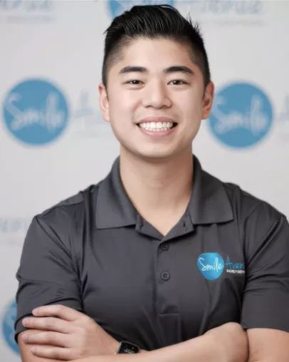 A smiling dentist with dark hair wearing a gray polo shirt featuring the Smile Avenue Family Dentistry logo stands in front of a backdrop with the same logo. The setting is likely at Smile Avenue in Katy, conveying professionalism and friendliness from the dental team.