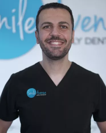 Smiling dentist in black scrubs with Smile Avenue Family Dentistry logo, standing in front of a dental clinic sign. Cypress & Katy family dentist offering emergency dental care.