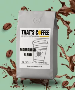A gray bag of Marrakesh Blend coffee with a label reading “THAT’S COFFEE” sits on a mint green background, surrounded by coffee splashes and beans—a perfect showcase for those who love discovering unique coffee blends.