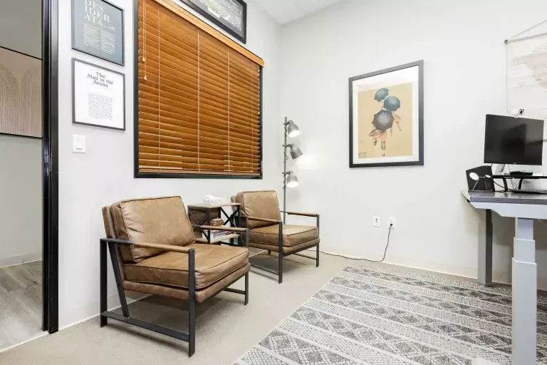 A modern office with two brown leather chairs, a small bookshelf, wall art, a window with wooden blinds, a patterned rug, and part of a desk with a computer monitor on the right.