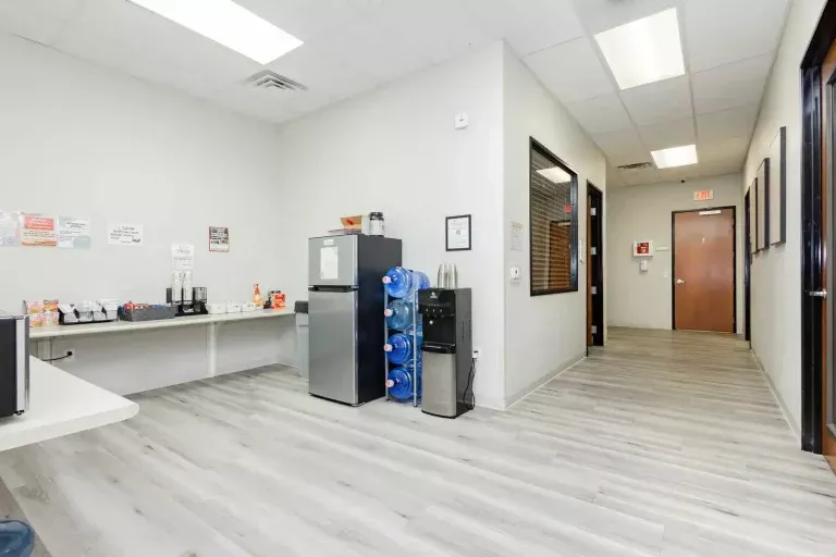 A modern office break room with a refrigerator, coffee makers, a water cooler with several blue water jugs, and a countertop with various supplies. The room has light gray floors and white walls with overhead lighting.