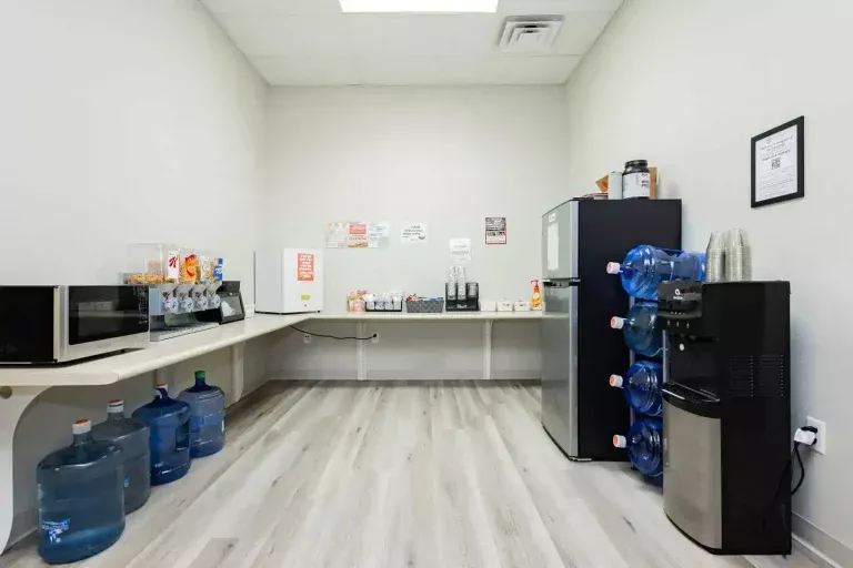 A workplace break room with microwaves, snack dispensers, a coffee station, a refrigerator, stacked water bottles, a water cooler, and disposable cups on light wood flooring and white walls.