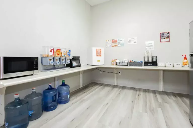 A break room with a microwave, cereal dispensers, a coffee maker, a mini fridge, cups, condiments, hand sanitizer, and several large water jugs on the floor, all against white walls and light wood flooring.