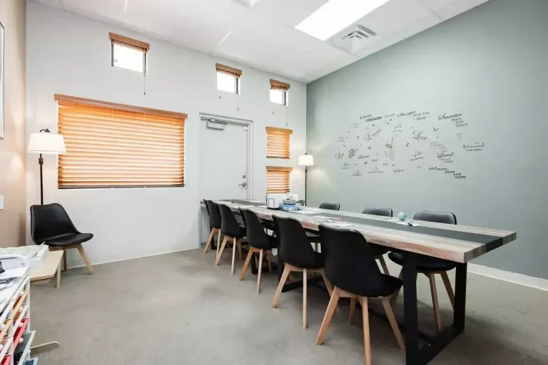 A modern conference room with a long wooden table, eight black chairs, a wall with writing and signatures, two lamps, and windows with wooden blinds. The room has neutral colors and a clean, minimalist design.