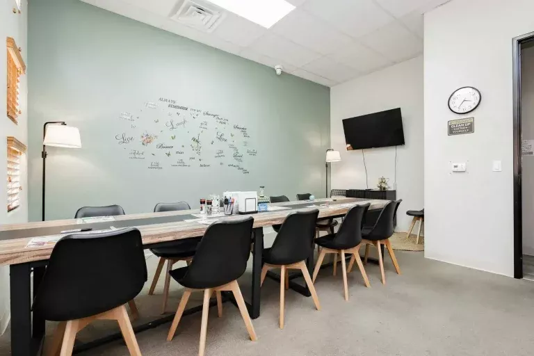 A modern conference room with a long table, ten black chairs, wall art with motivational words, a TV mounted on the wall, a clock, and two floor lamps. The room is well-lit and organized.