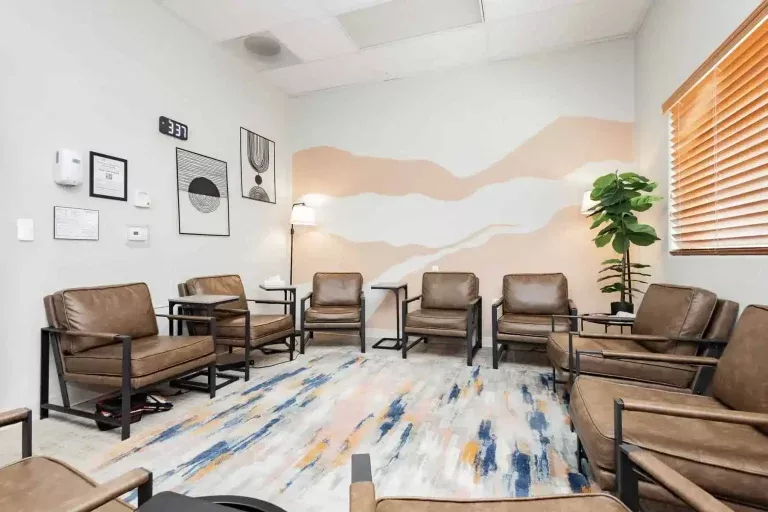 A modern waiting room with brown leather chairs arranged in a circle, a colorful rug, wall art, a potted plant, a tissue box, and a clock showing 3:37 on a white wall with abstract designs.