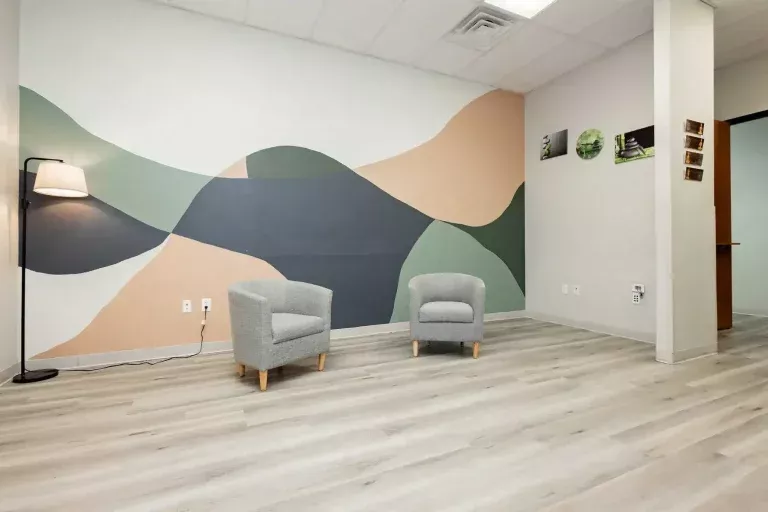 A modern room with light wood flooring, two gray armchairs, a floor lamp, and a wall mural featuring abstract shapes in green, beige, and black. Three small photos hang on a side wall. The space appears clean and minimalistic.