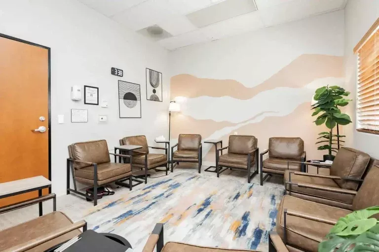 A modern waiting room with brown leather chairs arranged in a circle, a potted plant, wall art, a lamp, a wooden door, and a window with blinds. The floor has a colorful rug, and the walls are decorated with abstract designs.