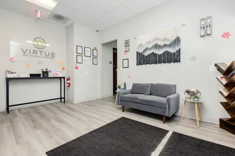 A clean, modern waiting room with a gray couch, small side table, wall art, and certificates on the walls. The floor is light wood, and a reception desk with the sign Virtue Recovery Center is in the background.