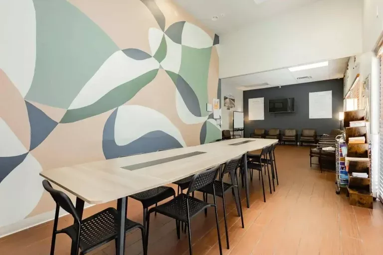 A modern meeting room with a long wooden table, black chairs, a large abstract mural on the wall, a TV, and a seating area with armchairs in the background. Natural light streams in from windows on the right.