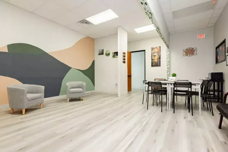 A modern office lounge with light wood flooring, abstract wall art, two gray armchairs, a white table with black chairs, wall decorations, and green plants. Fluorescent lights brighten the spacious, tidy room.