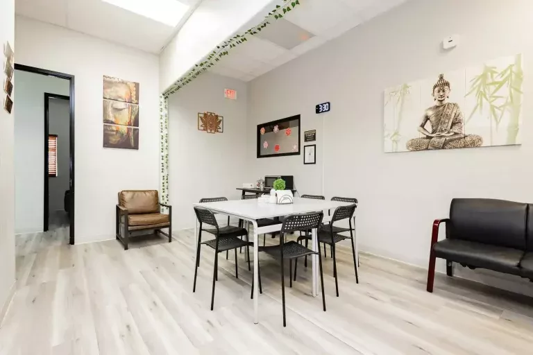 A bright, modern room with light wood floors, a white table surrounded by six black chairs, armchairs along the walls, wall art including a Buddha, and greenery on the ceiling. A clock, certificates, and decor are visible.