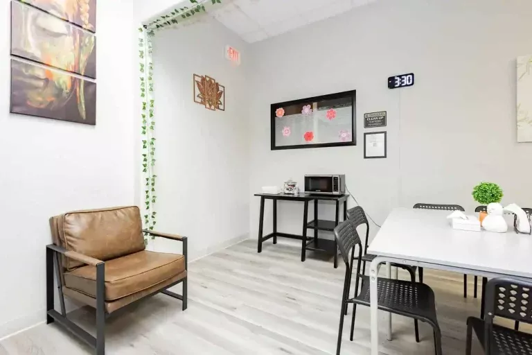 A bright break room with a tan armchair, a table with black chairs, a microwave on a side table, wall art, a digital clock showing 3:30, and green vines decorating one corner.