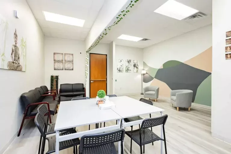 A modern waiting room with a white table, black chairs, black sofas, gray armchairs, wall art, plants, and a geometric accent wall. The space is brightly lit with overhead lights.