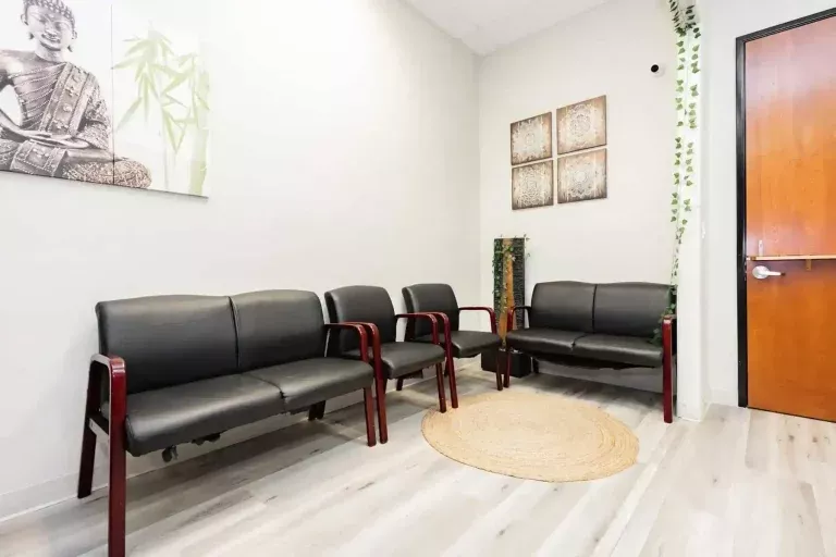 A bright waiting room with black chairs, a round beige rug, wall art, a wooden door, and green plants, featuring light-colored floors and white walls.