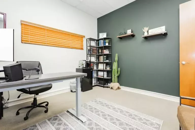 A modern office with a desk, chair, laptop, bookshelf, two floating shelves, a cactus plant, a window with blinds, and a patterned rug on a beige carpeted floor.
