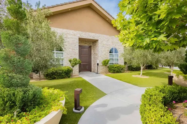 A modern house with a stone facade, tall front door, and arched window, surrounded by manicured bushes, trees, and a curved sidewalk on a neatly trimmed lawn under a sunny sky.