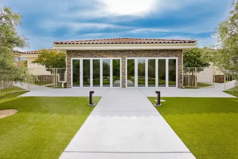 A modern building with a stone facade, large glass doors, and a tiled roof, surrounded by green artificial grass, trees, and a concrete walkway leading to the entrance under a partly cloudy sky.