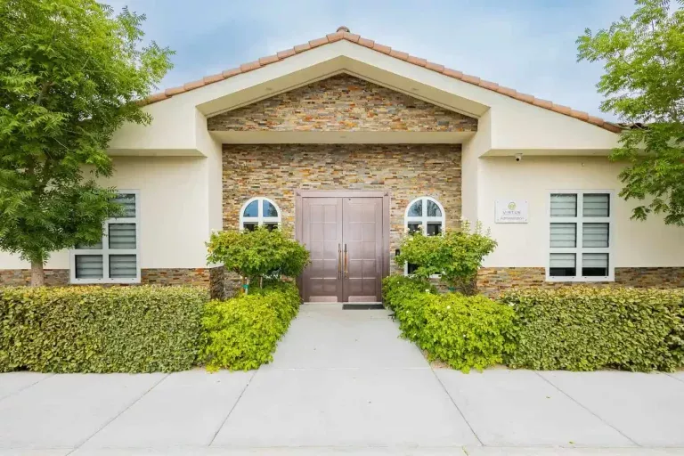 A modern building with stone and cream exterior, arched windows, double front doors, trimmed hedges, and two trees on either side of the entrance under a cloudy sky.