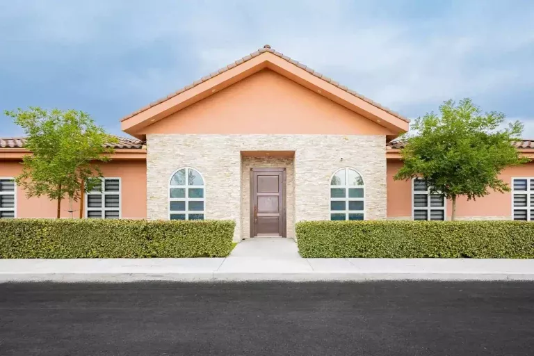 Single-story peach-colored building with a central brown door, arched windows on each side, trimmed hedges, two small trees, and a paved street in front under a partly cloudy sky.