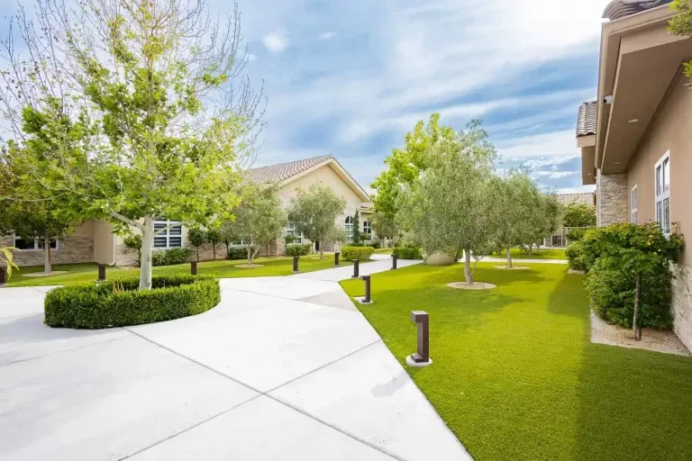 A landscaped courtyard with green grass, trees, shrubs, and pathways leading to light-colored residential buildings under a partly cloudy sky.