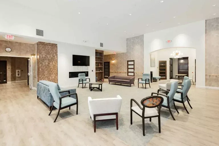 A spacious, bright lounge area with various chairs and sofas arranged in a circle, light wood flooring, neutral walls, a large TV, bookshelves, and modern decor.