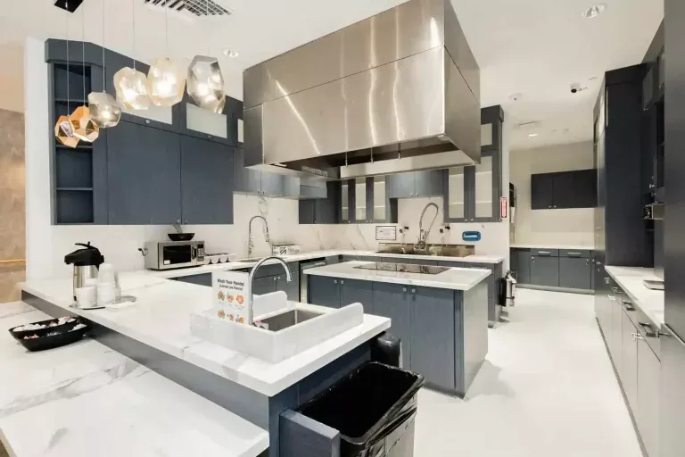 Modern kitchen with blue cabinets, white countertops, stainless steel appliances, large range hood, double sinks, pendant lights, and coffee makers on the counter. The space is clean, bright, and spacious.