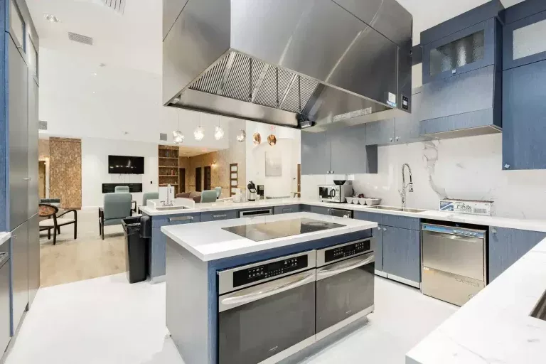 Modern kitchen with blue cabinets, stainless steel appliances, a large vent hood, white countertops, and an island with built-in ovens. The space opens into a dining and living area with light wood flooring.