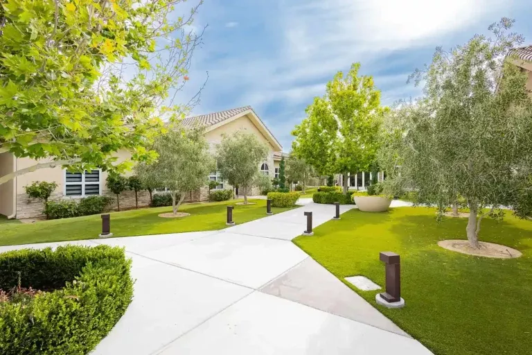 A sunny courtyard with neatly trimmed grass, trees, shrubs, and a wide concrete walkway leading to beige buildings with tile roofs and windows. Modern pathway lights line the walkway, and the sky is bright with light clouds.