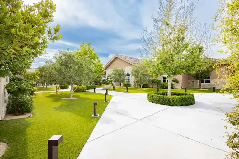 A wide, curved driveway leads to a single-story house surrounded by manicured green lawns, trees, and neatly trimmed hedges under a partly cloudy sky.