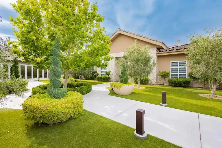 A well-maintained yard with trimmed bushes, trees, and green grass surrounds a beige house with white-trimmed windows, stone accents, and a winding white walkway under a blue sky.