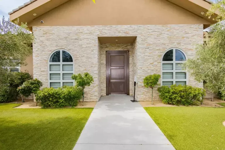 A modern house entrance with a large wooden door, arched windows on both sides, light stone exterior, manicured shrubs, and a straight concrete walkway surrounded by green grass.