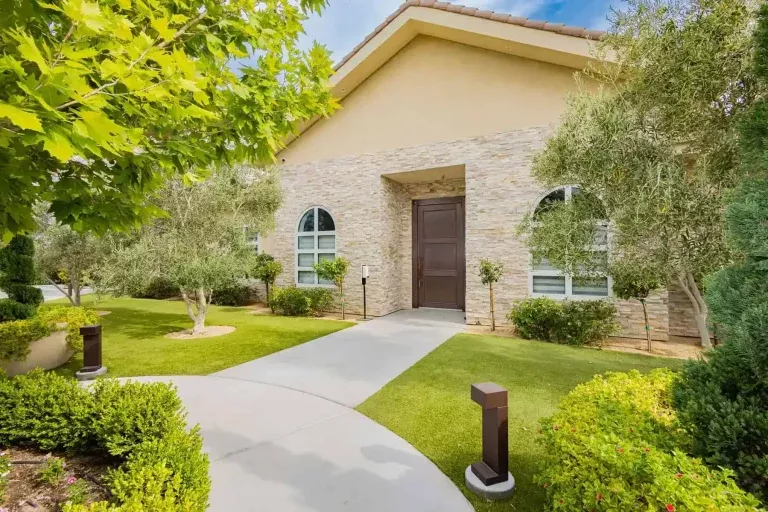 A modern single-story house with a light stone facade, arched windows, and a dark wooden front door, surrounded by neatly trimmed grass, lush green bushes, and small trees along a curved walkway.
