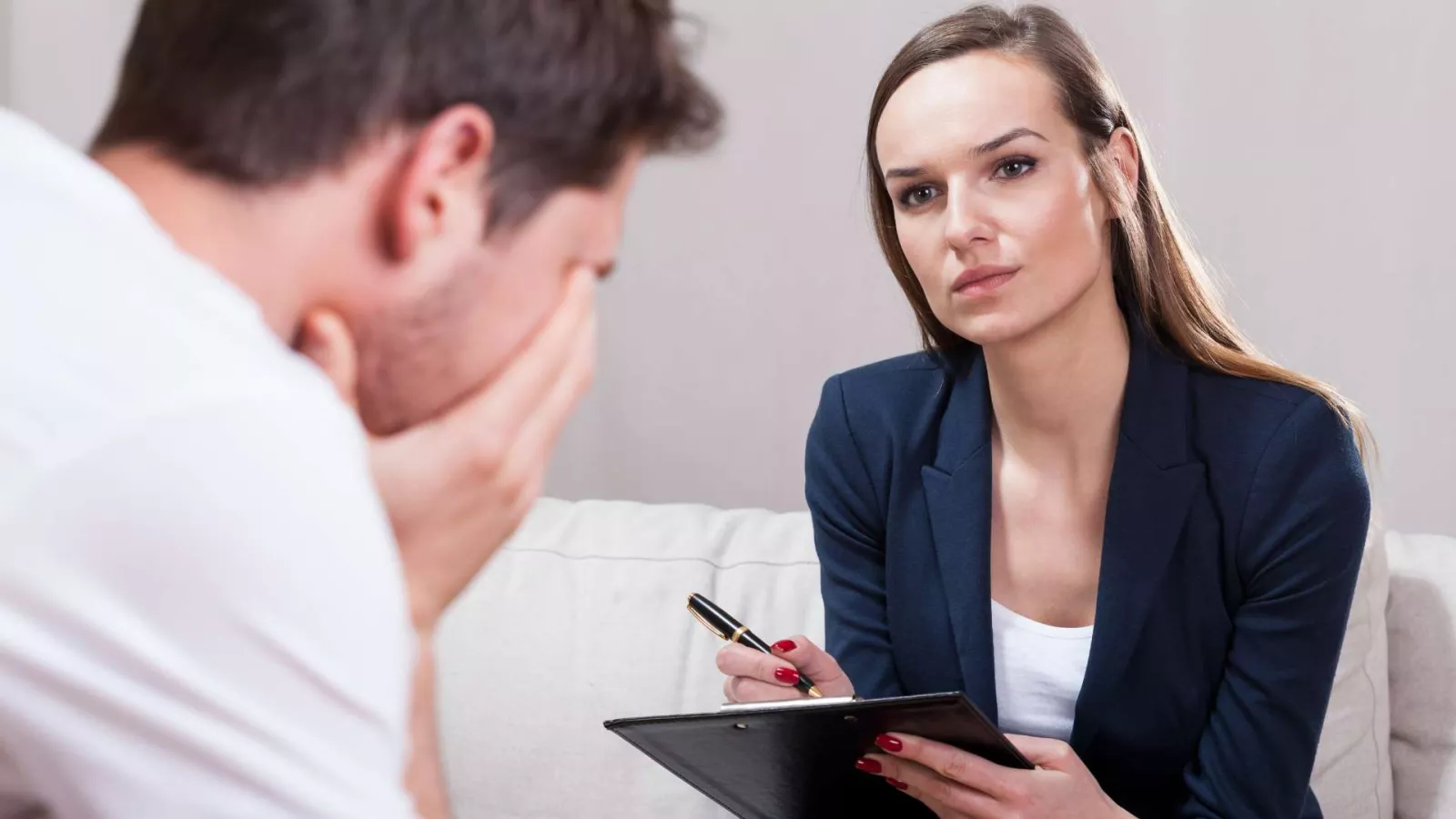 A woman holding a clipboard and pen listens attentively to a man who is sitting with his head in his hands, appearing distressed. They are seated on a couch in what looks like a counseling or therapy session.