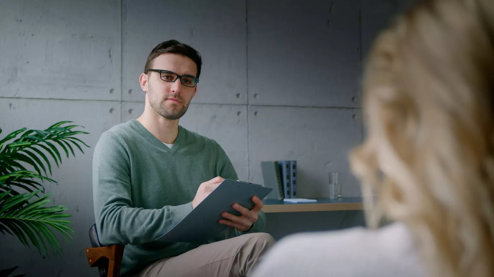 A man wearing glasses and a green sweater sits with a clipboard, attentively listening to a woman whose back is to the camera during a conversation in a modern, minimalistic office setting.