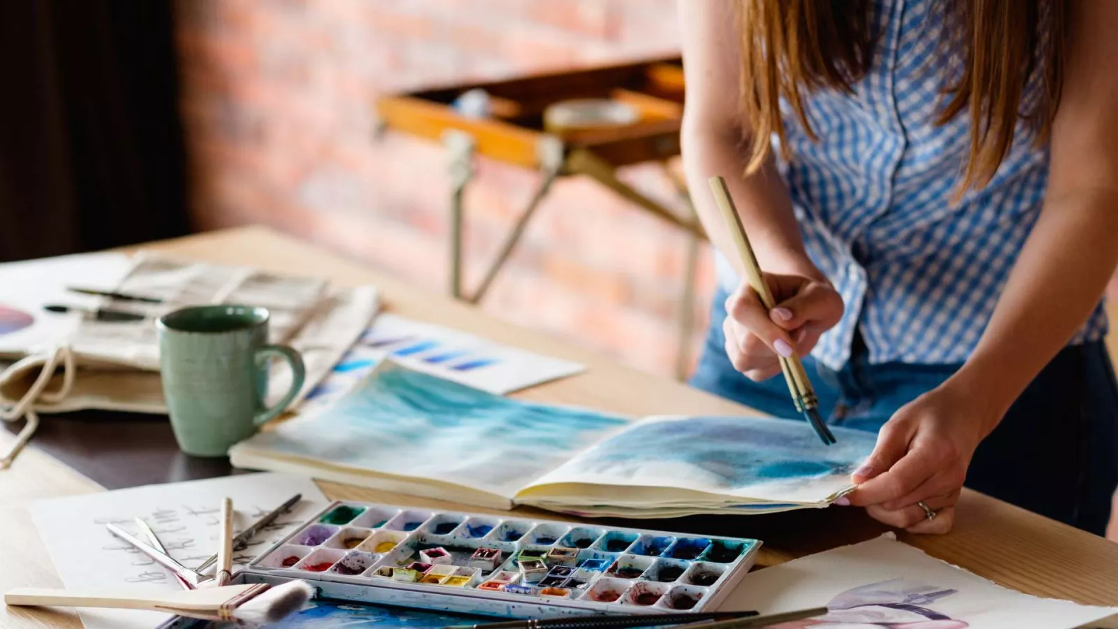 A person in a blue checkered shirt paints in a sketchbook with a brush, using watercolors. Art supplies, a mug, and papers are spread out on a wooden table under natural light.