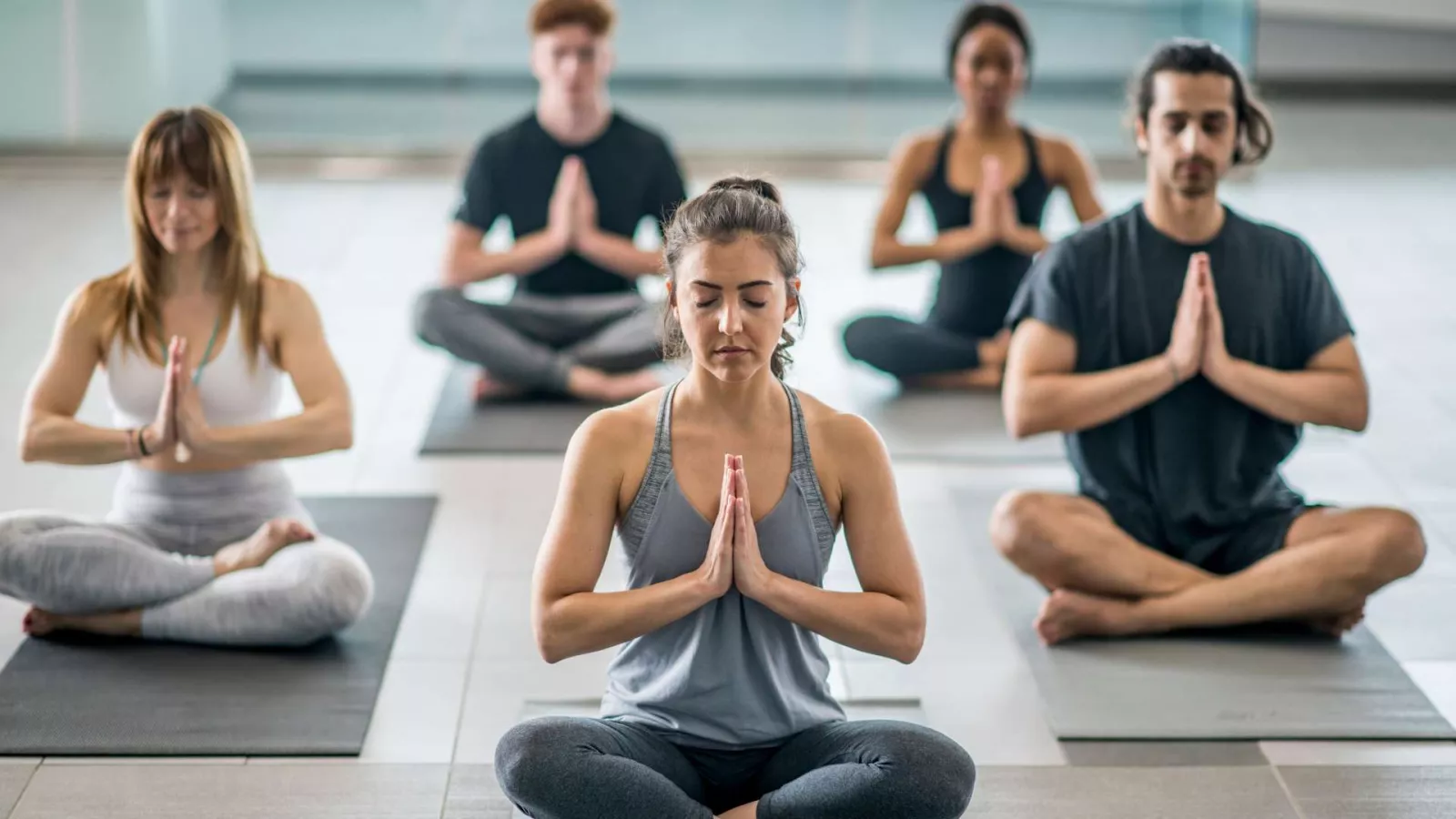 Five people sit cross-legged on yoga mats indoors, eyes closed and hands pressed together in a prayer position, practicing meditation or yoga in a calm, bright studio.