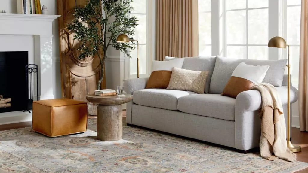 A cozy seating area in a home featuring a sofa with pillows and a throw blanket, a lamp, an ottoman, a small table, and other decor. Products are from Bassett Furniture.